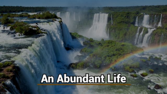 Aerial view of expansive waterfalls cascading into a river surrounded by lush greenery, with a rainbow arching over the mist. Text overlay states &ldquo;An Abundant Life.&rdquo; Opens a new window with the video.