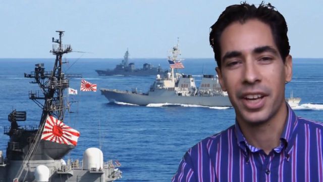 A man in a striped purple shirt speaking in front of a digitally added background depicting a naval scene with warships on a calm ocean, one displaying a large rising sun flag. Opens a new window with the video.