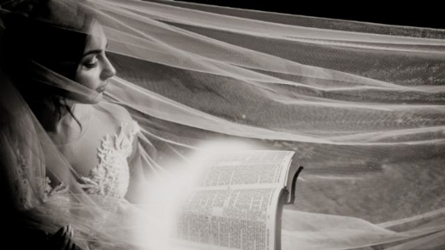 A black and white image depicting a woman with a serene expression, draped in a translucent veil, gazing intently at an open book emitting a bright, glowing light. The setting evokes a sense of mystical enlightenment. Opens a new window with the video.