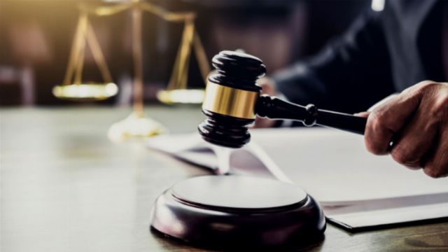 A judge in a black robe about to use a gavel, with a digital tablet and a document on the table in front of them. Scales of justice are visible in the softly lit background. Opens a new window with the video.