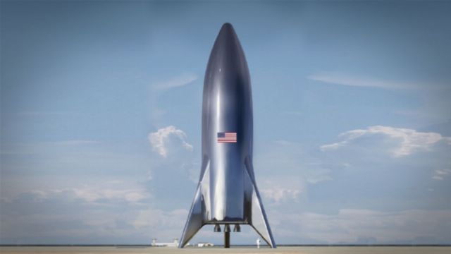 A large, sleek rocket stands on a launch pad under a clear blue sky with scattered clouds. The rocket is adorned with a United States flag. Opens a new window with the video.