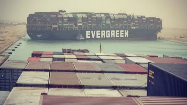 A large cargo ship named EVERGREEN is positioned diagonally across a narrow canal, surrounded by sandy banks under a hazy sky. In the foreground, numerous colorful shipping containers are stacked at a port. Opens a new window with the video.