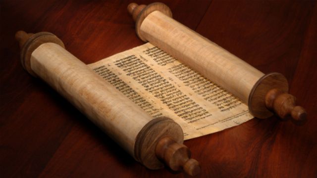 An ancient scroll open on a wooden table, with Hebrew text visible, possibly a sacred document. Opens a new window with the video.