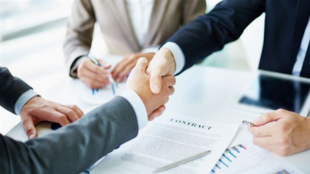 A close-up photo showing two individuals engaging in a firm handshake over a table with business documents and financial charts spread out, implying the closure of a successful negotiation. Another person appears to be taking notes, participating in the agreement process. Opens a new window with the video.