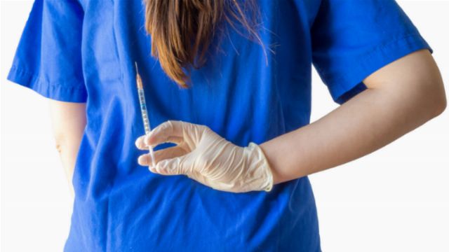 A healthcare worker, dressed in a blue uniform, holds a syringe in a gloved hand, ready for administration. The person's face is unseen, emphasizing the act of medication preparation. Opens a new window with the video.