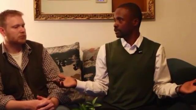 Two men seated on a sofa engaged in a discussion, with one man extending his hands in an animated gesture. A world map and ornate frame decorate the background. Opens a new window with the video.