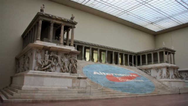 A large classical sculpture reproducing an ancient monument displayed in a museum. The sculpture features a grand staircase flanked by a series of dynamic figures in relief, depicting historical or mythological events. A partially visible informational blue banner with white and red text covers some lower steps leading up to the monument, under a vast glass ceiling that diffuses soft natural light onto the exhibit. Opens a new window with the video.
