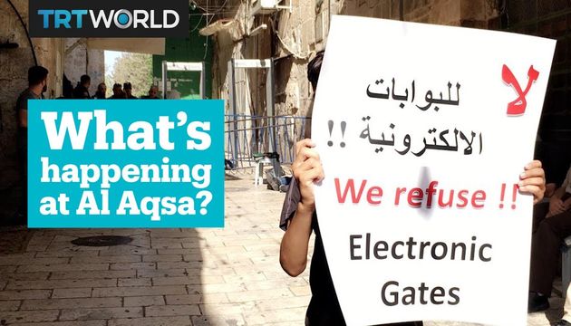 A person in a street holding two signs, one in English and one in Arabic. The English sign reads, &ldquo;We refuse!! Electronic Gates.&rdquo; To the left, a graphic element with the text &ldquo;What&rsquo;s happening at Al Aqsa?&rdquo; is visible. The scene features Middle Eastern architecture in the background, conveying a public demonstration or protest atmosphere. Opens a new window with the video.