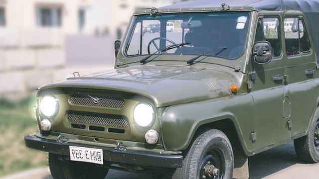 An olive green vintage off-road vehicle parked outdoors, featuring a distinctive classic design with round headlights and a visible license plate. Opens a new window with the video.