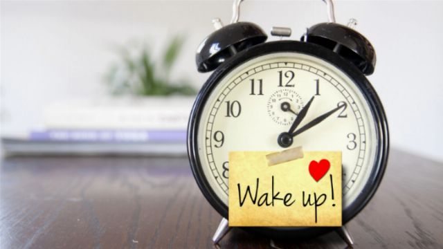 A traditional black alarm clock with a sticky note attached, reading &ldquo;Wake up!&rdquo; adorned with a red heart, set on a wooden surface. A blurred book and a plant in the background suggest a calm, domestic setting. Opens a new window with the video.
