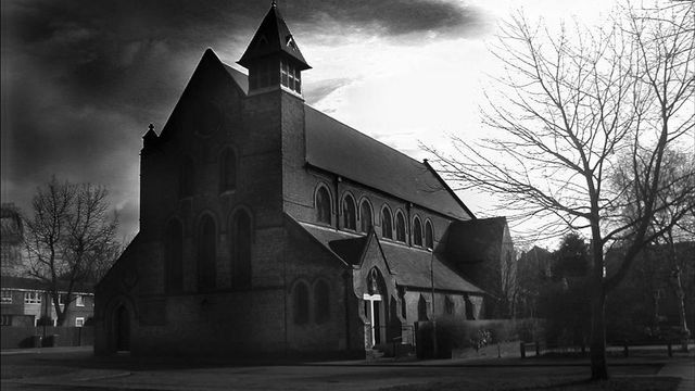 Black and white image of a Gothic-style church under a dramatically cloudy sky, with leafless trees to the right and a crucifix visible near the entrance. Opens a new window with the video.