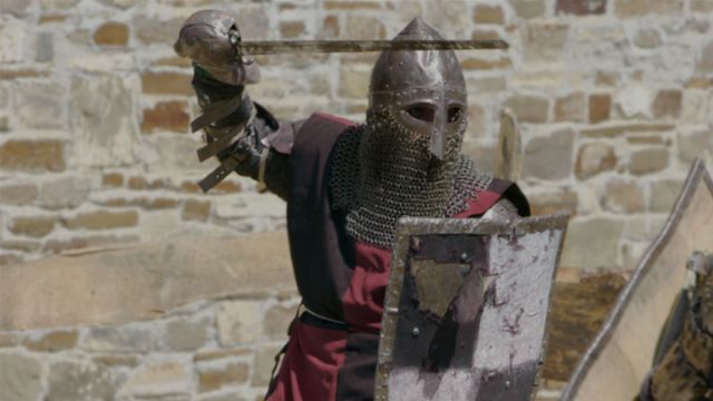 A knight in traditional armor, complete with a chainmail hood and helmet, holds a sword in a striking posture behind a weathered shield, against a backdrop of an ancient stone wall. Opens a new window with the video.