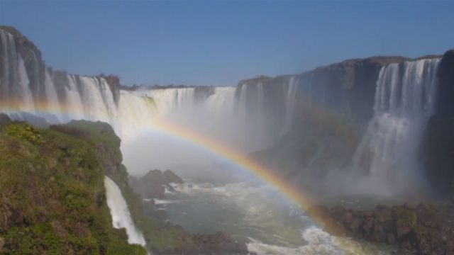 A majestic view of a large waterfall system, with multiple waterfalls plunging into a river. A vibrant rainbow arches across the misty spray in the foreground, under a clear blue sky. Opens a new window with the video.