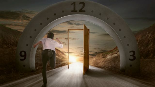 A man is running towards an open door framed by a large circular structure marked with numbers from a clock, positioned on a rural road leading to a horizon where the sun sets dramatically. Opens a new window with the video.