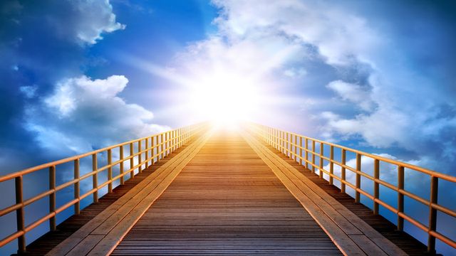 A wooden bridge leading towards a luminous sun, which forms a bright backdrop against a sky scattered with clouds. Opens a new window with the video.
