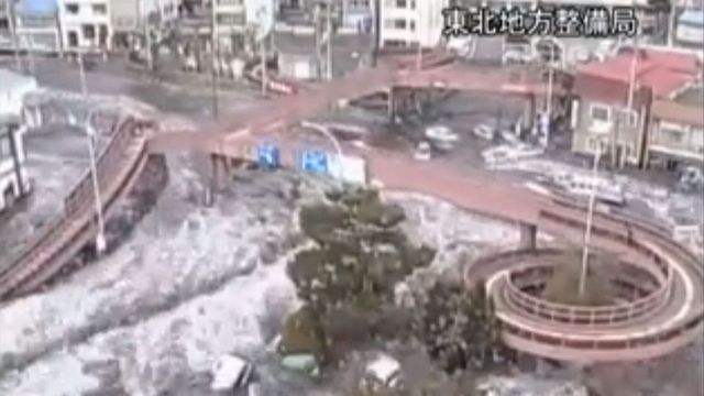Aerial view of an urban area with roads and buildings partially covered by snow, alongside a clear view of a circular structure and green trees. Opens a new window with the video.
