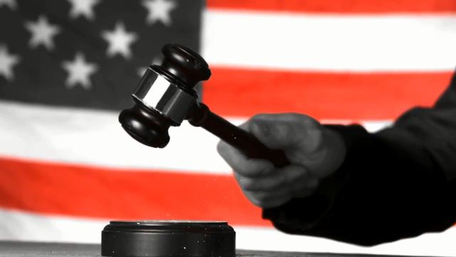 A monochrome image selectively colored depicting a hand striking a gavel down, set against a blurred backdrop of the American flag. Opens a new window with the video.