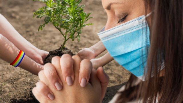 A young woman wearing a surgical mask and two other individuals plant a small tree together in soil. The woman's eyes are closed, and one of the hands assisting her displays a rainbow-colored bracelet. Opens a new window with the video.