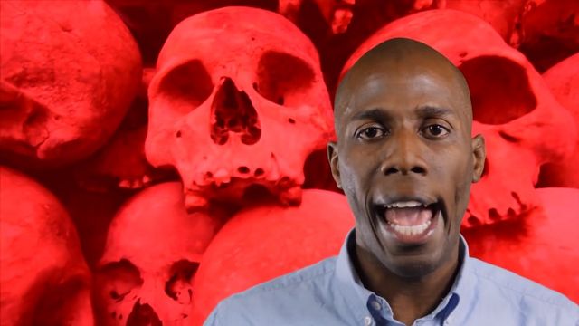 An African American man, appearing surprised or speaking emphatically, is standing in front of a vividly red background adorned with multiple skull images. Opens a new window with the video.