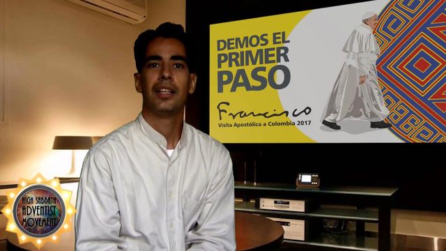 A man in a white shirt is seated in front of a poster displaying a graphical illustration depicting a figure in white robes and a slogan related to a 2017 event in Colombia. A small digital clock sits on a desk beside him, as well as a logo related to a movement features prominently in the bottom left of the image. Opens a new window with the video.