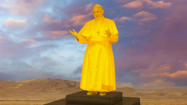 A statue of a religious figure, arrayed in a golden robe, stands on a pedestal with a serene expression, gesturing subtly with both hands. The background features a dynamic sky with hues of blue and pink above a barren desert landscape. Opens a new window with the video.