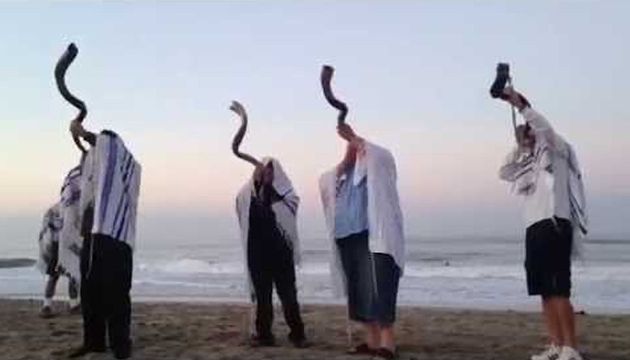 Four individuals on a beach at dusk, each holding a shofar, a traditional horn used in ceremonies, to their mouths. They wear prayer shawls over their heads, facing the sea as waves gently lap the shore. Opens a new window with the video.