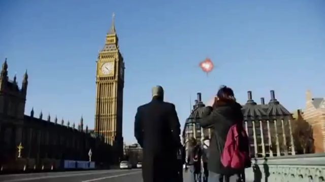 A group of people on a bridge, observing a bright unidentified aerial phenomenon in the shape of a heart above them, with the iconic Big Ben and the British Parliament buildings in the backdrop under a clear blue sky. Opens a new window with the video.