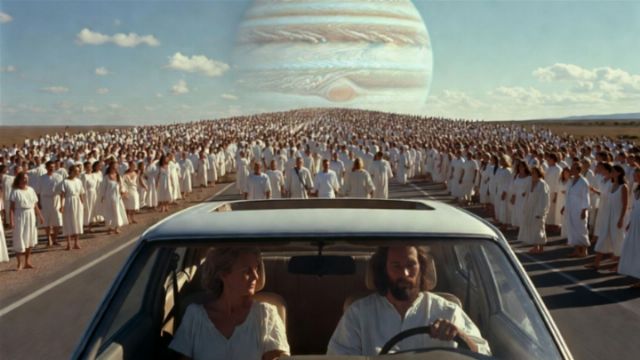 A car is parked on a road with two occupants sitting inside. In the foreground, a man is driving while a woman is seated beside him. In the background, a large crowd of people dressed in white garments is gathered, appearing to march toward a massive image of the planet Jupiter in the sky. Opens a new window with the video.