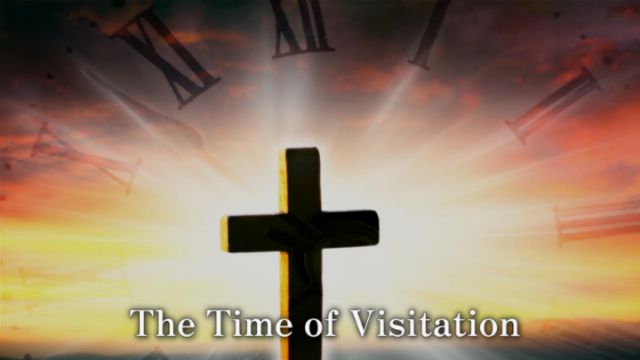A silhouette of a cross stands centered against a vibrant, cloudy sky, illuminated by rays of light. Abstract shapes resembling ancient time marking symbols hover faintly in the background, surrounding the scene. The text &ldquo;The Time of Visitation&rdquo; is displayed at the bottom. Opens a new window with the video.