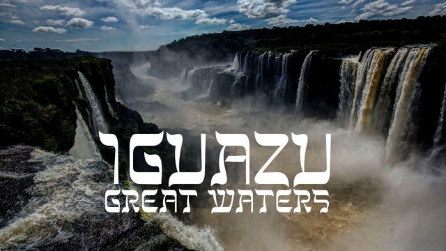 Majestic view of Iguazu Falls, showcasing cascading waterfalls plunging powerfully into mist-covered depths under a partly cloudy sky. Bold text overlay reads &ldquo;IGUAZU GREAT WATERS&rdquo;. Opens a new window with the video.