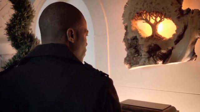 A man with a shaved head, viewed from behind, gazes at an illuminated sculptural relief depicting a celestial theme reminiscent of the Mazzaroth, mounted on a white arched wall. The artwork radiates a warm, golden glow, and a subtle wreath decorates the arched entry. Opens a new window with the video.