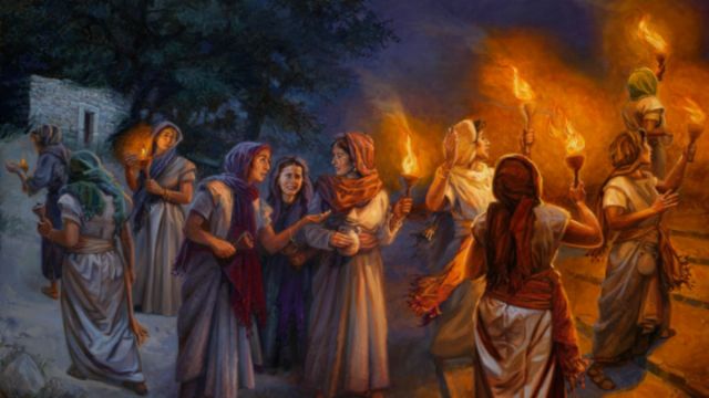 A gathering of women in ancient attire walking in a night scene, holding torches that illuminate their faces. Some women are engaged in conversation, while others are looking toward a distant house. The background features trees and a stone building. Opens a new window with the video.