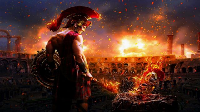 A muscular warrior in traditional Roman attire, wielding a sword and shield, stands over a fiery pit in a grand ancient arena. The sky is illuminated with flames and smoke, creating a dramatic atmosphere as the warrior prepares for battle. Opens a new window with the video.