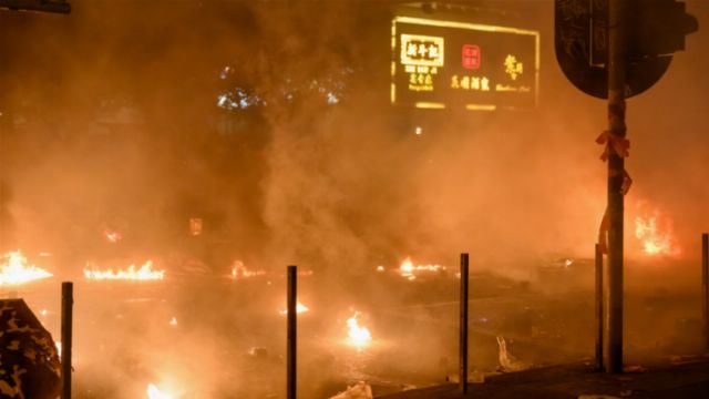 A nocturnal city scene illuminated by multiple small fires scattered across a street. Smoke obscures parts of the background, which includes an illuminated street sign displaying characters in a non-Latin script. Opens a new window with the video.