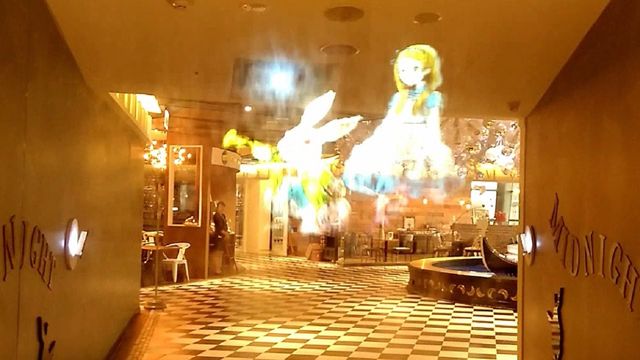Interior of a brightly lit venue with a checkered floor and holographic projection of a girl and a rabbit on the wall. A faint glow resembling celestial constellations illuminates the space, contributing to a mystical atmosphere reminiscent of the Mazzaroth. Opens a new window with the video.