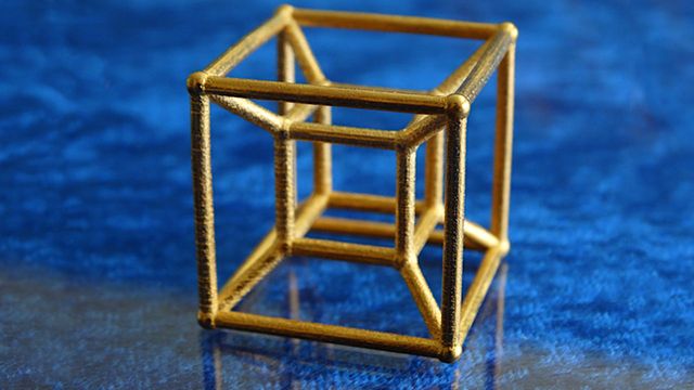 A golden geometric sculpture resembling a three-dimensional cube structure, displayed on a blue textured surface. Opens a new window with the video.
