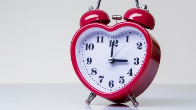 A heart-shaped red alarm clock with a white face displaying the time set to three minutes past three, standing against a plain light background. Opens a new window with the video.