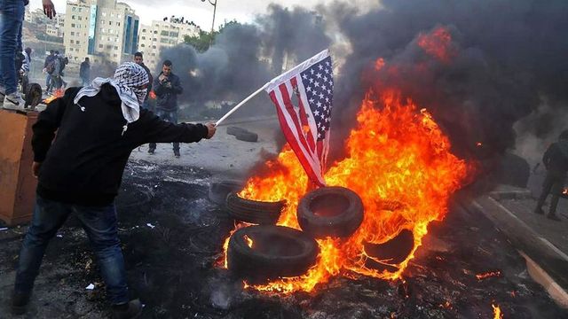 A person with a covered face holding an American flag over a fire made from burning tires in an urban setting with buildings visible in the background. Smoke fills the air. Opens a new window with the video.