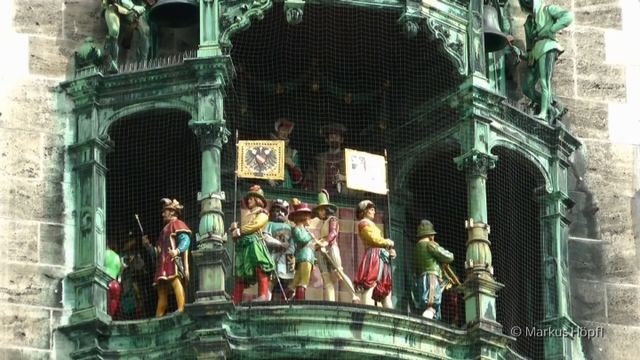 Mechanical figures in historical costumes perform on an ornate clock tower's balcony, surrounded by sculptural details and stone architecture. The central figure holding a shield adorned with a starry representation, possibly referring to the Mazzaroth, is flanked by others with musical instruments and regal attire. Opens a new window with the video.