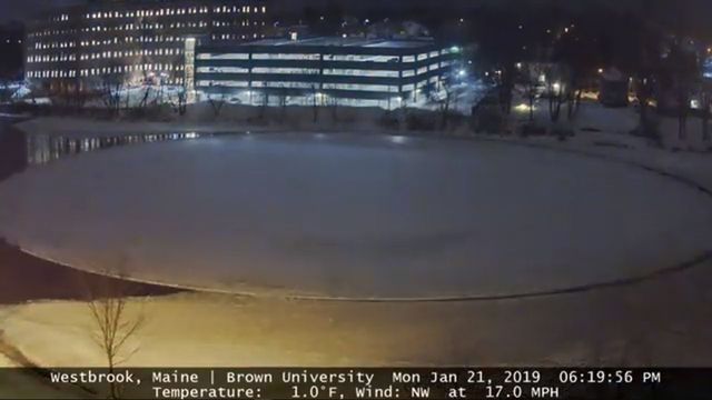 A winter night scene showing a frozen pond in the foreground with a softly lit multi-story building in the background. The sky appears clear, reflecting a typical cold evening under a starry firmament. The image includes a timestamp and weather data: Westbrook, Maine, Brown University, with a temperature of 1.0&deg;F and a northwest wind at 17.0 MPH, dated January 21, 2019 at 06:19:56 PM. Opens a new window with the video.