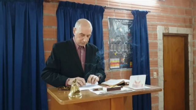 A man standing at a wooden podium in a room with a brick wall and blue curtains. He is reviewing notes and books, including one that appears open. A small bell is also on the podium. A chart resembling a celestial map is posted in the background, potentially depicting the Mazzaroth. Opens a new window with the video.