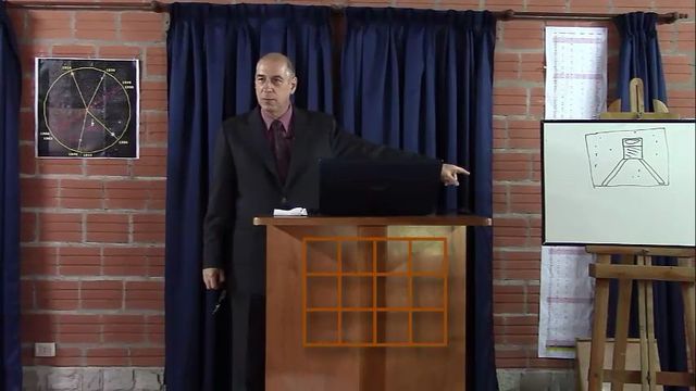 A man stands at a podium in a room, gesturing towards a laptop on the podium, with a framed image of the Mazzaroth and an illustration of a chemical flask on an easel to his side. Opens a new window with the video.
