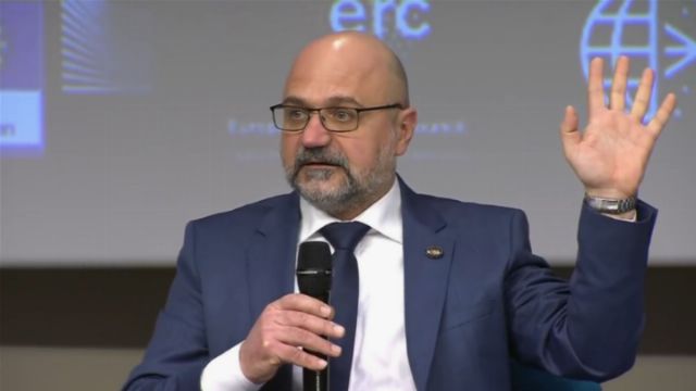 A man with a beard dressed in a suit holds a microphone and gestures with his left hand while speaking at a conference. The background displays a logo with a globe and the initials &ldquo;ERC.&rdquo; Opens a new window with the video.
